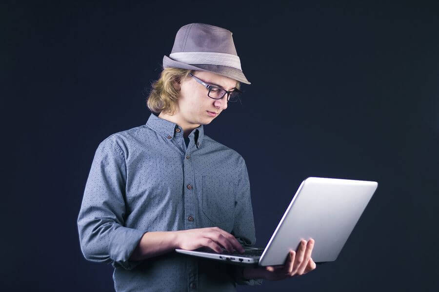 Lehrlingsportal.at Eine Person mit Brille und grauem Fedora tippt auf einem silbernen Laptop vor einem dunklen Hintergrund.