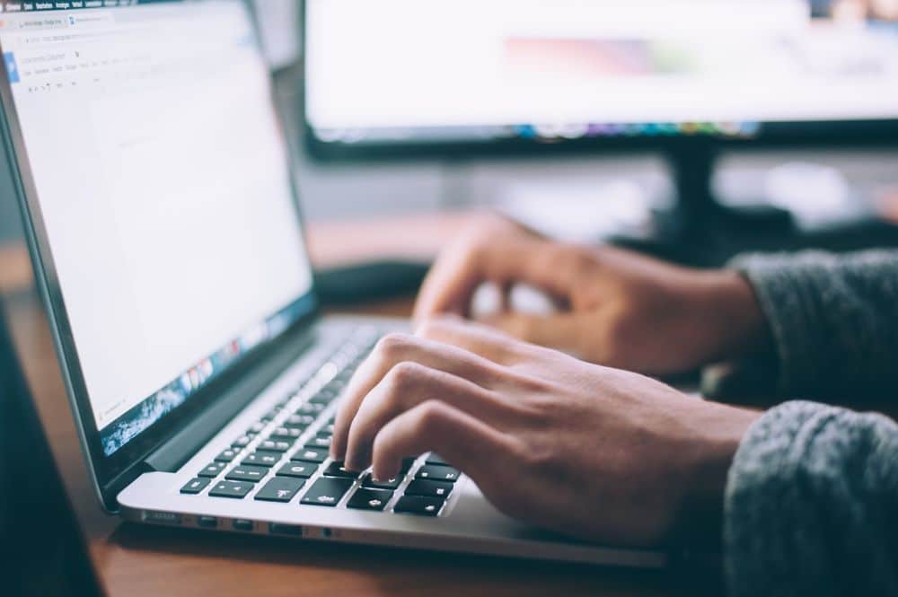 Lehrlingsportal.at Person tippt auf einem Laptop mit zwei Monitoren im Hintergrund.