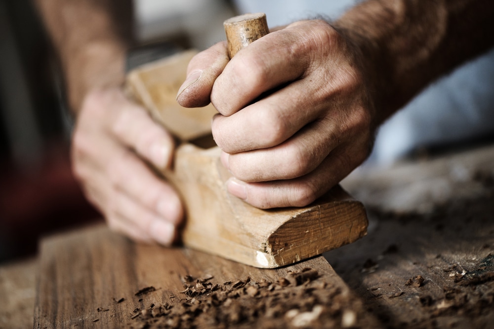 Lehrlingsportal.at Hände verwenden einen Holzhobel, um eine Holzoberfläche zu glätten, wobei Holzspäne verstreut herumliegen.