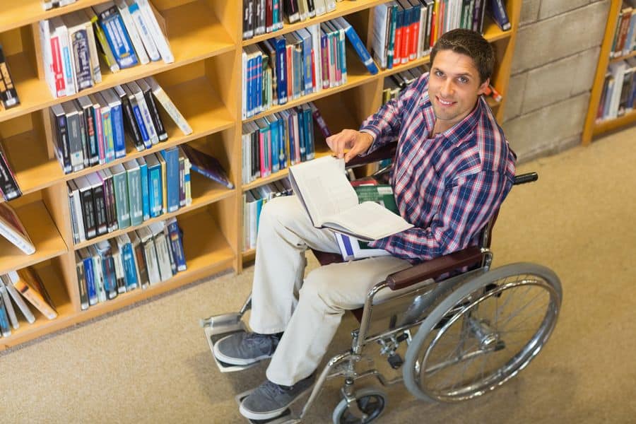 Lehrlingsportal.at Eine Person im Rollstuhl hält ein offenes Buch und lächelt in einer Bibliothek. Im Hintergrund sind Regale voller Bücher zu sehen.