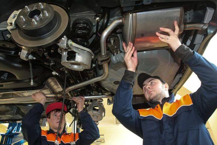 Lehrlingsportal.at Zwei Mechaniker arbeiten in blauen und orangefarbenen Uniformen in einer Werkstatt unter einem Auto und prüfen und justieren Teile.