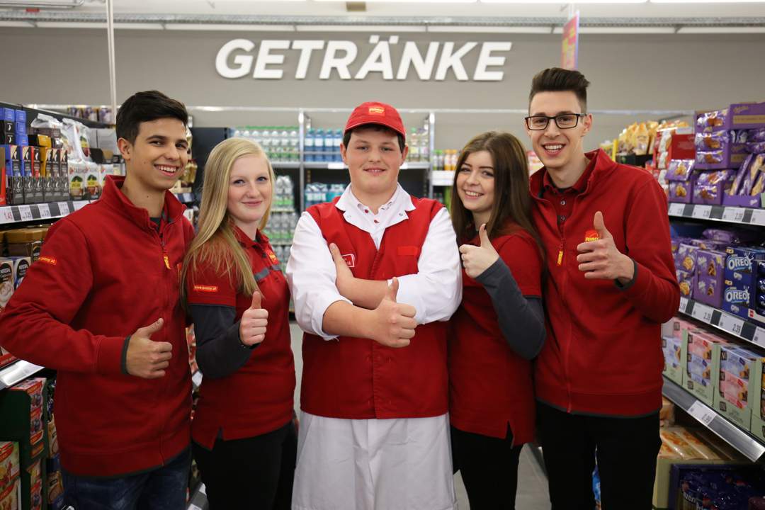 Lehrlingsportal.at Fünf Supermarktmitarbeiter in roten Uniformen stehen in einem Gang mit Regalen voller Produkte und strecken den Daumen nach oben.