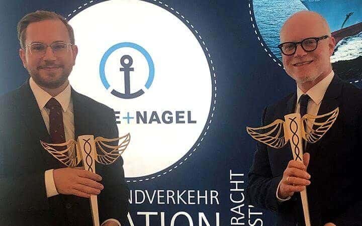 Lehrlingsportal.at Zwei Männer in Anzügen halten hölzerne Caduceus-Modelle in der Hand und stehen vor einem runden Logo mit einem Ankersymbol und dem Text „Kuehne+Nagel“.