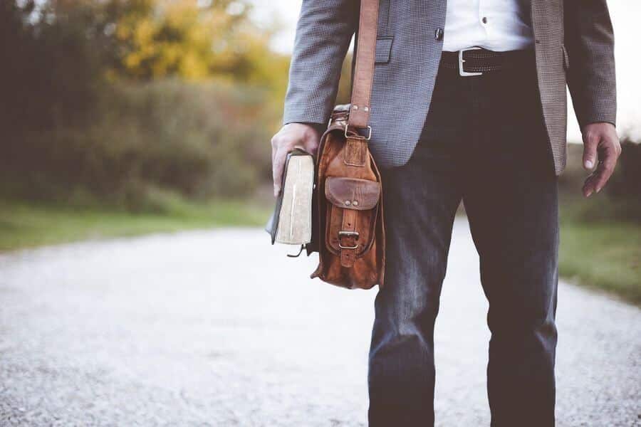 Lehrlingsportal.at Eine Person im Blazer hält ein Buch und eine braune Ledertasche und steht auf einem Kiesweg mit viel Grün im Hintergrund.