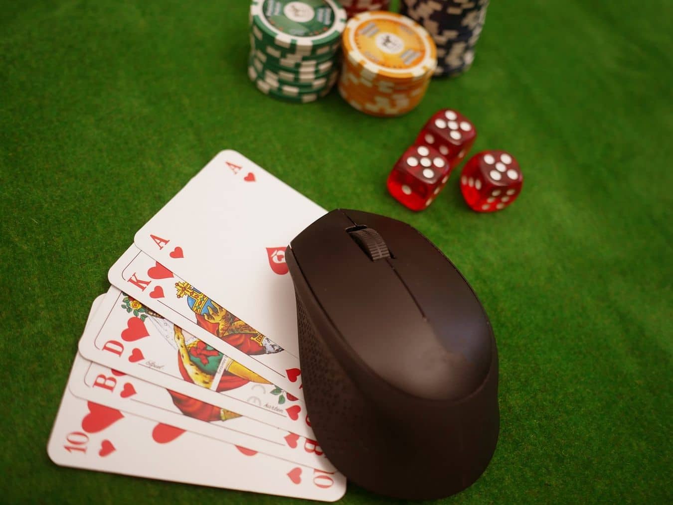 Lehrlingsportal.at Eine Computermaus ruht auf Spielkarten, darunter eine Herz Zehn, ein Herz Bube, eine Herz Dame, ein Herz König und ein Herz Ass, neben Pokerchips und zwei roten Würfeln auf einer grünen Filzoberfläche.