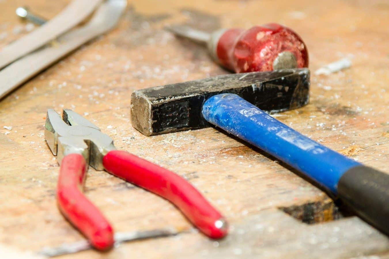 Lehrlingsportal.at Auf einer hölzernen Werkbank liegen eine Zange mit rotem Griff, ein Hammer mit schwarzem Kopf und blauem Griff sowie ein Schraubenzieher mit rotem Griff.