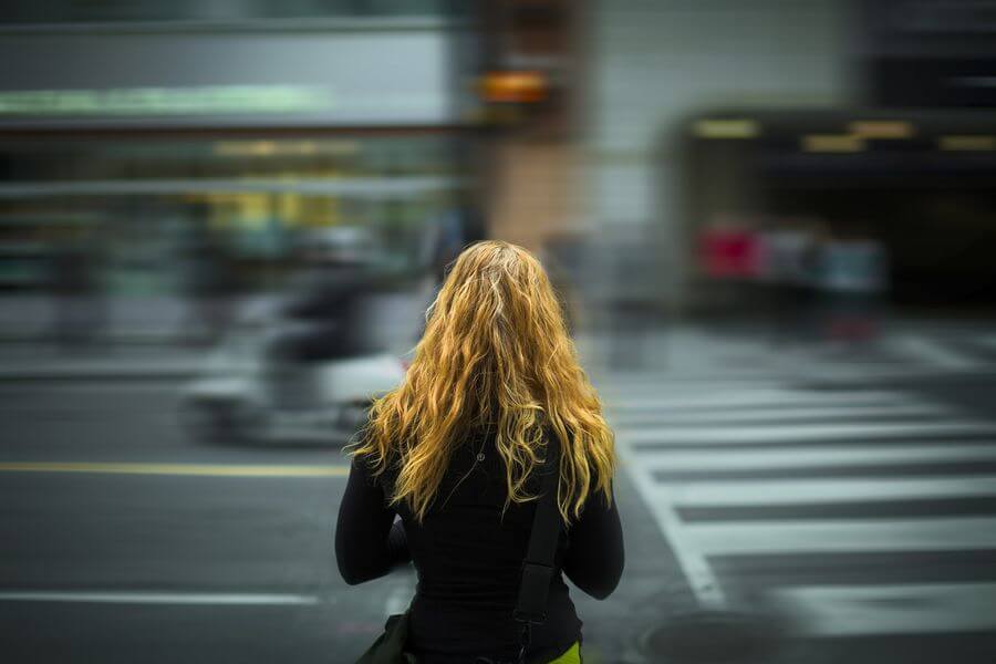 Lehrlingsportal.at Eine Person mit langen blonden Haaren steht an einem viel befahrenen Zebrastreifen in der Stadt und blickt auf die verschwommene Sicht auf Verkehr und Gebäude.