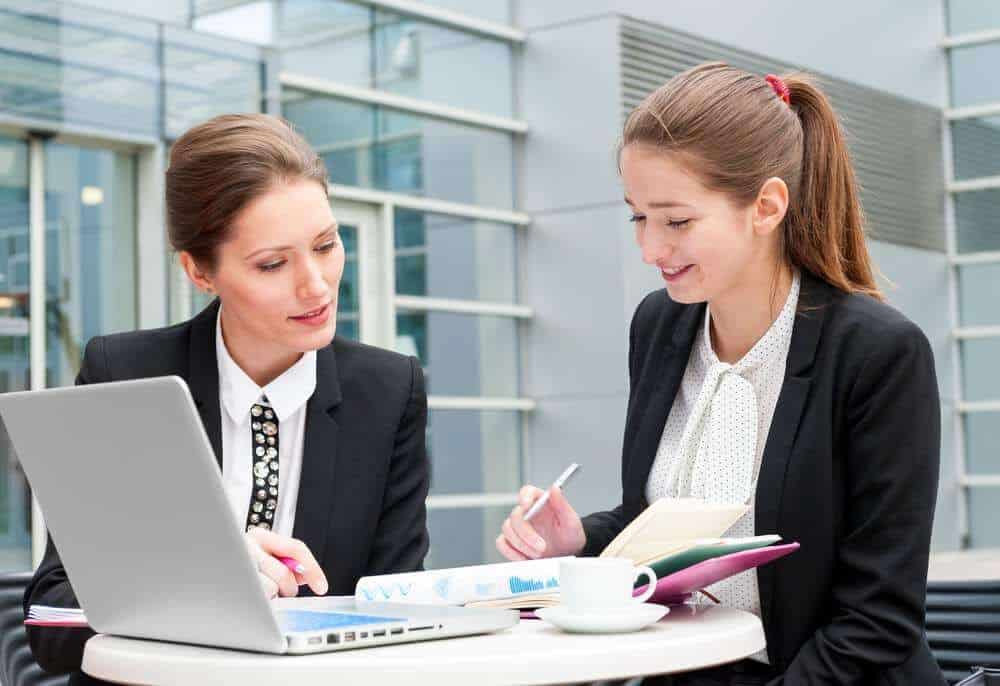 Lehrlingsportal.at Zwei Frauen in Business-Kleidung sitzen mit einem Laptop und Notizbüchern an einem Tisch und unterhalten sich vor einem Bürogebäude.