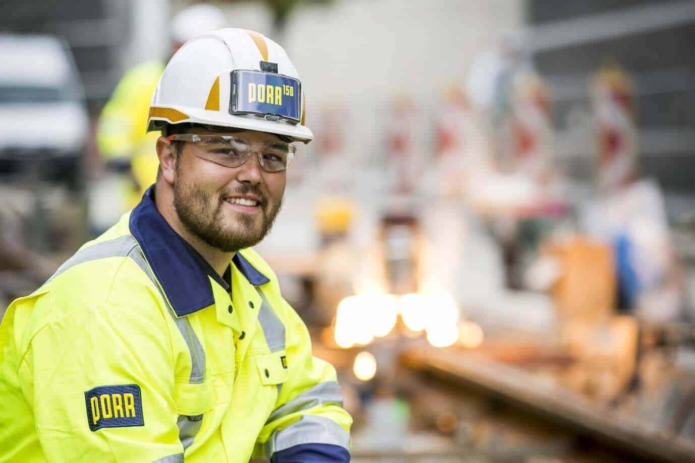 Lehrlingsportal.at Ein Bauarbeiter mit Schutzhelm und Warnweste lächelt in die Kamera. Im Hintergrund ist eine unscharfe Baustelle zu sehen.