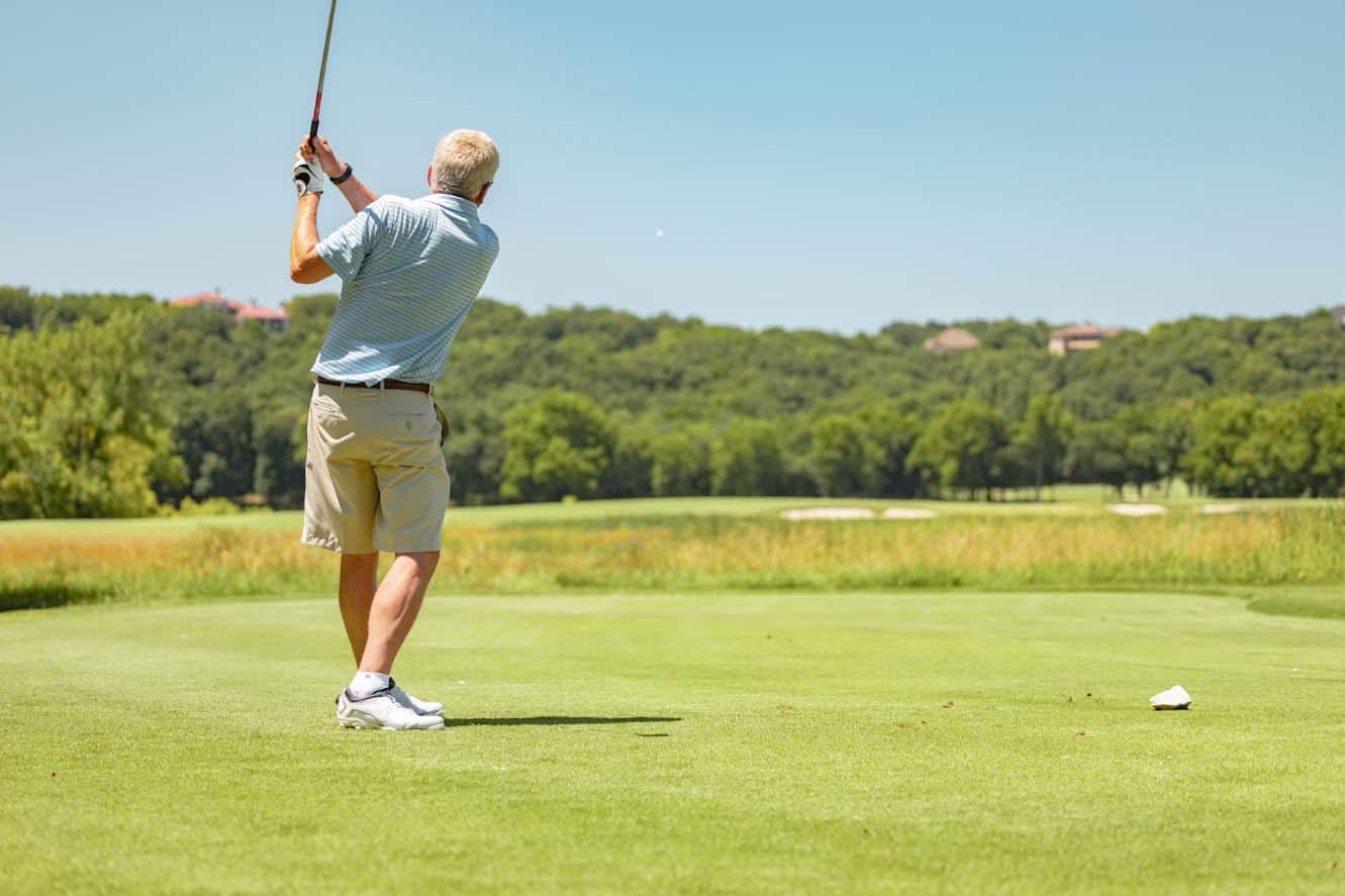 Lehrlingsportal.at Eine Person in gestreiftem Hemd und Shorts spielt auf einem grünen Golfplatz und schwingt unter einem klaren blauen Himmel ihren Schläger.