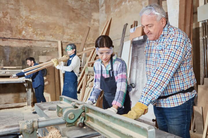 Lehrlingsportal.at Gruppenarbeit in einer Werkstatt. Ein Mann und eine Frau bedienen eine Tischkreissäge, während andere im Hintergrund Bretter tragen und zuschauen. Alle tragen Schutzausrüstung.