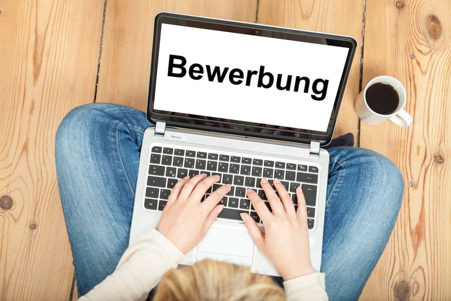 Lehrlingsportal.at Person, die einen Laptop mit dem Wort „Bewerbung“ verwendet und auf einem Holzboden sitzt, neben ihr eine Tasse Kaffee.