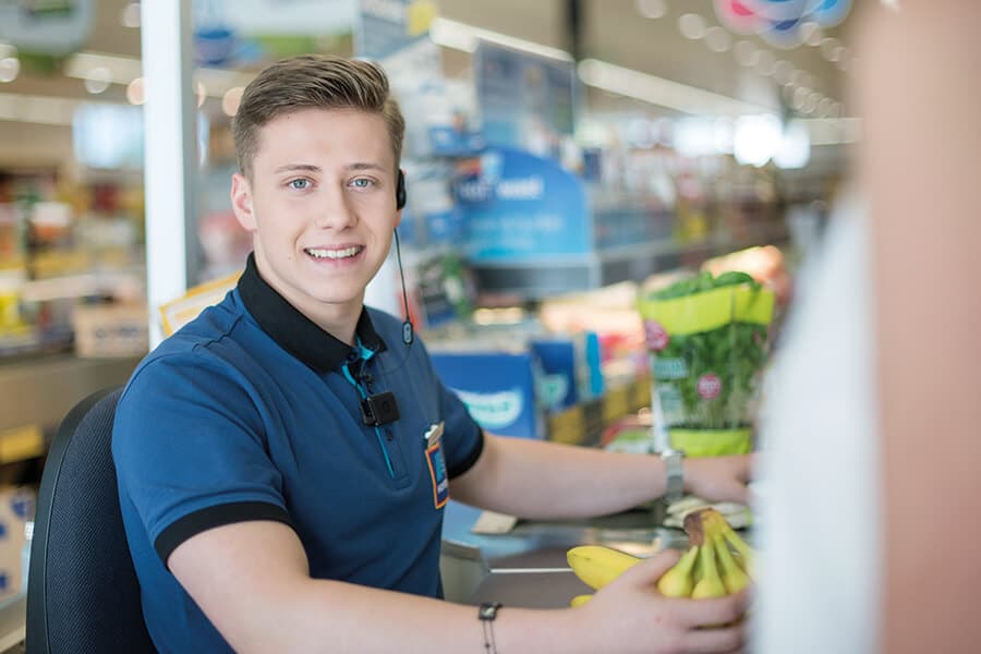 Lehrlingsportal.at Eine Kassiererin in blauer Uniform scannt Bananen an einer Kasse in einem Lebensmittelgeschäft.