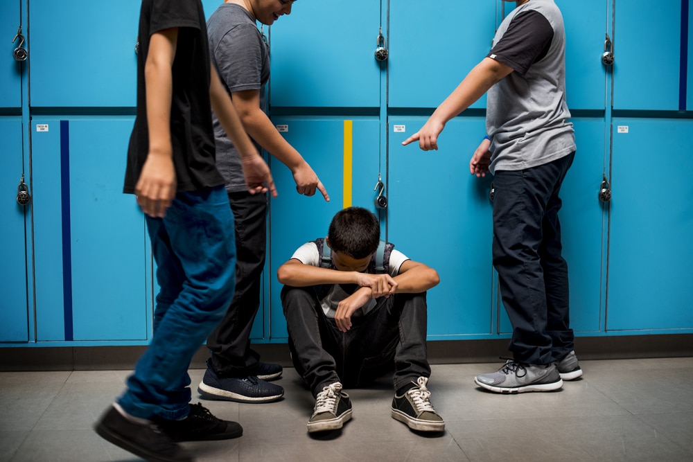 Lehrlingsportal.at Ein sitzender Schüler mit gesenktem Kopf ist vor blauen Schließfächern von drei stehenden Mitschülern umgeben, die auf ihn zeigen.