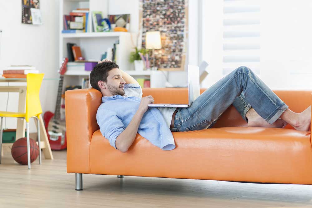 Lehrlingsportal.at Eine Person liegt mit einem Laptop auf einem orangefarbenen Sofa. Im Zimmer stehen eine Gitarre, ein Basketball, Bücher und ein gelber Stuhl.