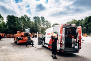 Lehrlingsportal.at Ein Arbeiter mit Schutzhelm steht neben einem offenen Lieferwagen mit der Aufschrift „HANSA-FLEX“. In der Nähe parkt auf einem Industriegelände im Freien ein orangefarbener Gabelstapler. Im Hintergrund sind weitere Geräte zu sehen.