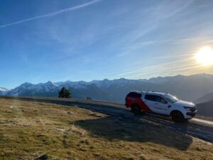 Lehrlingsportal.at Ein Rettungsfahrzeug auf einer Bergstraße mit schneebedeckten Gipfeln im Hintergrund unter einem klaren blauen Himmel.