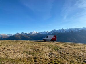 Lehrlingsportal.at Weißer und roter Lieferwagen, geparkt auf einer grasbewachsenen Hügelkuppe mit Panoramablick auf schneebedeckte Berge unter einem klaren blauen Himmel.