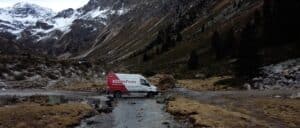 Lehrlingsportal.at Ein Lieferwagen mit der Aufschrift „Hansa Flex“ parkt in einer Bergregion auf einem steinigen Pfad, umgeben von schneebedeckten Gipfeln und spärlicher Vegetation.