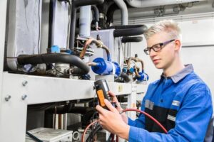 blonder Lehrling mit Brille und blauer Arbeitskleidung prüft Maschine