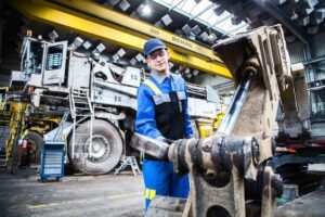 Lehrling in Arbeitskleidung befesteigt Schaufel am Bagger