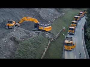 Lehrlingsportal.at Ein Bagger führt Arbeiten am Hang aus, während mehrere Lastwagen eine Bergstraße säumen und ein Arbeiter in der Nähe den Verkehr regelt.