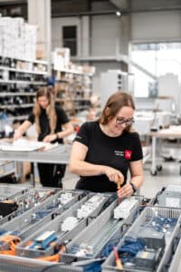 Lehrlingsportal.at Zwei Frauen arbeiten in einer Industrieanlage und montieren elektrische Komponenten an einem Tisch. Im Hintergrund sind Regale mit Kisten zu sehen.