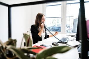 Lehrlingsportal.at Eine Frau im schwarzen Blazer telefoniert mit einem Festnetztelefon und blickt dabei auf einen Computermonitor in einem hellen Büro. Im Vordergrund steht eine Pflanze.