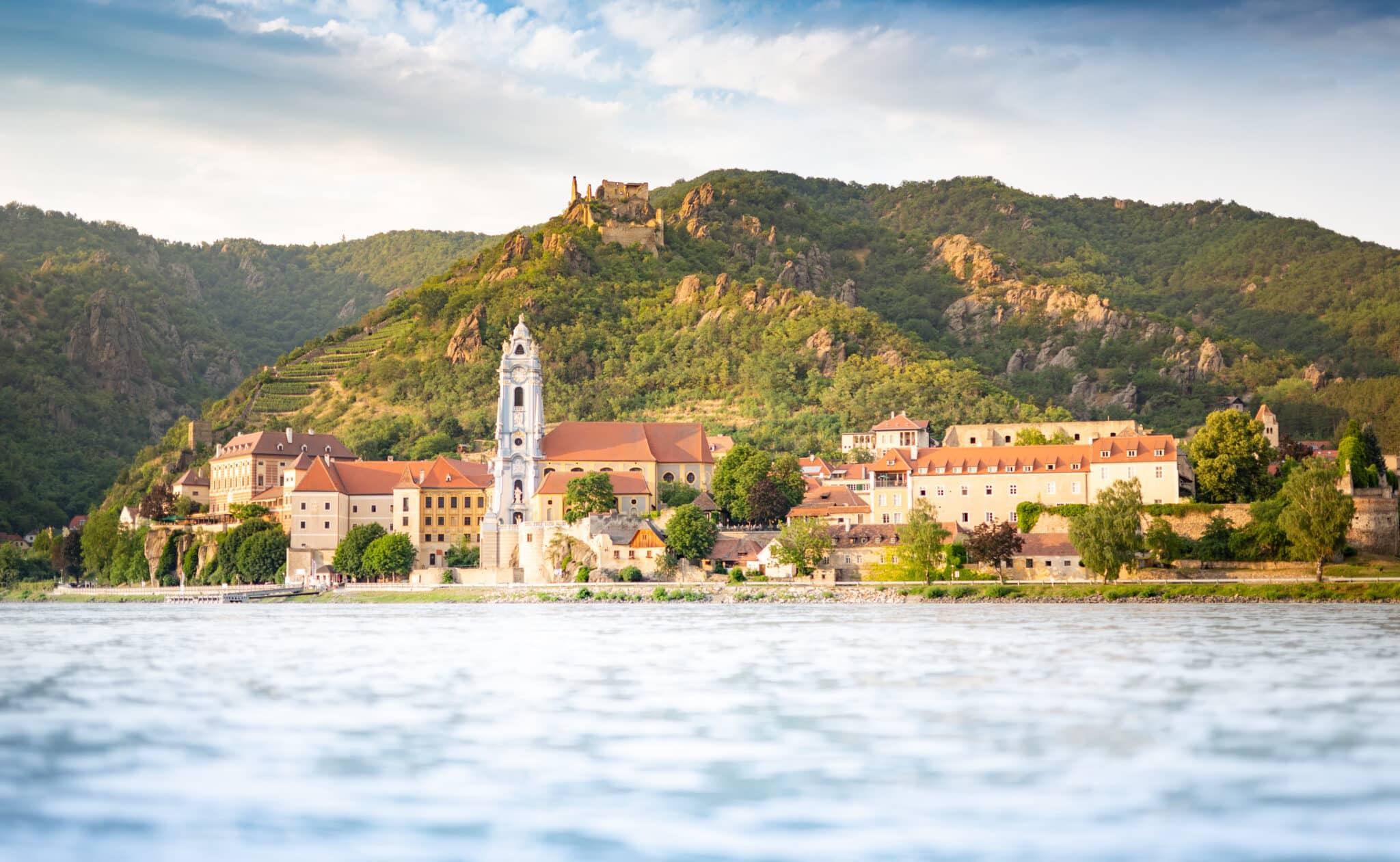 Lehrlingsportal.at Malerischer Blick auf eine historische Stadt mit einem markanten blauen Kirchturm, eingebettet zwischen grünen Hügeln und einem fließenden Fluss unter einem teilweise bewölkten Himmel.