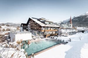 Lehrlingsportal.at Alpenhotel mit großem Außenpool, umgeben von schneebedeckten Bergen und Gebäuden, unter klarem Himmel.