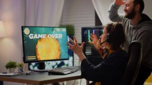 Lehrlingsportal.at Eine Person mit Headset sitzt an einem Computer, auf dem ein „Game Over“-Bildschirm angezeigt wird, während eine andere Person mit erhobener Hand dahinter steht.