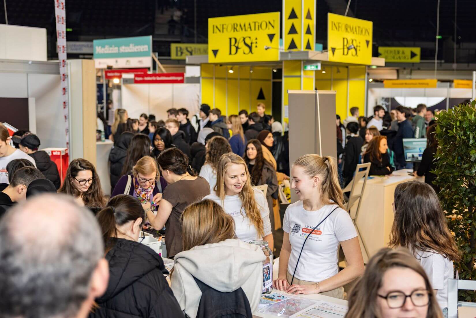 Lehrlingsportal.at Menschen besuchen eine gut besuchte Karrieremesse in einer großen Halle, im Hintergrund Informationsstände und Beschilderungen.