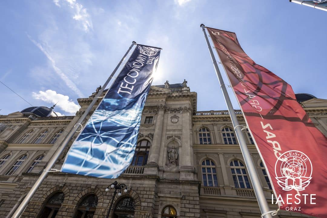 Lehrlingsportal.at Blick nach oben auf ein historisches Gebäude mit zwei vertikalen Bannern im Vordergrund, eines blau und eines rot, vor einem klaren Himmel.